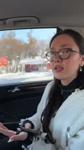 A woman is in the backseat of a car, moving past a wintery landscape, as evidenced by snow-covered trees visible through the window. She is wearing glasses, a black top under a white cardigan, and dangling earrings. She is holding a small, dark object in her left hand, possibly a phone or wallet. The car interior is dark, with hints of wood trim and a grey blanket visible beside her. She appears to be speaking or singing along to music that is playing in the car.