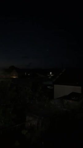 The video is a still shot of an outdoor scene at night. The sky is dark, with some faint clouds visible. In the background, there are several houses and buildings, some of which are illuminated by artificial lights. A street light is on and illuminating a road. Trees and foliage are prominent in the foreground, obscuring some of the view of the buildings. The overall impression is of a quiet, dimly lit residential area.