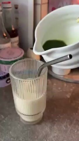 A person is pouring green liquid into a glass filled with white liquid. A metal straw sits in the glass. The green liquid is being poured from a white pitcher. The liquid is being poured at a medium-fast pace. In the background are bottles and containers of other liquids and powders. The liquid being poured is adding to the liquid already in the glass. The liquid in the glass is becoming a lighter shade of green as more liquid is poured. The countertops and surfaces are grey.
