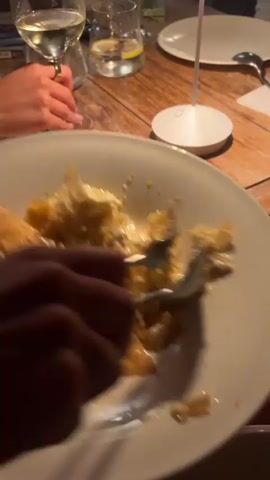 A close-up shot focuses on a person's hands using a fork and spoon to eat from a large white plate. The plate contains a dessert with flaky pastry and a creamy sauce. In the background, a wooden table is visible, along with other plates, wine glasses, and what appears to be a cell phone. The lighting is dim, suggesting an indoor setting like a restaurant. The person's clothing, a white dress with a gold necklace, is partially visible.
