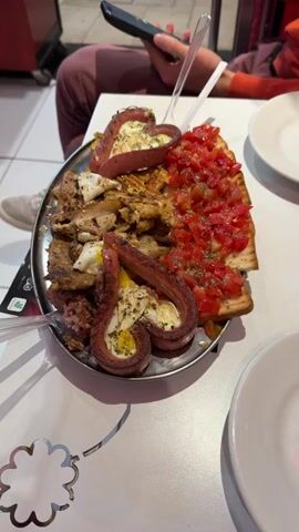 Here's a detailed description of the video:

The video shows a close-up of a large silver platter of food. On the platter, there is a mix of items including:

*   Grilled meat, which appears to be a mix of chicken and pork, cut into bite-sized pieces.
*   Sliced tomatoes, finely diced, piled on top of toasted bread slices.
*   Two sausages curved into a heart-like shape with fried eggs inside.

The platter is sitting on a white table with a floral pattern cut into the surface. In the background, two people are visible. One is holding a phone, and there are clear plastic forks sticking into the food. There are also two empty white plates on the table. The overall setting seems to be in a restaurant or casual dining environment.