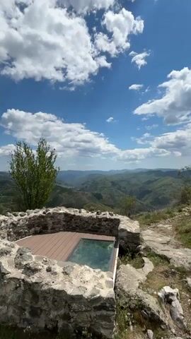 The video shows a stone structure with a wooden deck and glass bottom, overlooking a green mountainous landscape under a partly cloudy sky. A young woman walks into frame, pauses briefly holding a mobile phone, then walks away. The camera pans from the structure to show the broad landscape, trees, and the cloudy sky.