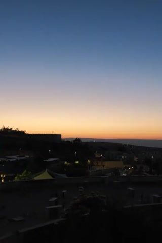 The video shows a panoramic view of a town at dusk. The sky is a gradient of deep blue at the top, fading to orange and then a soft yellow near the horizon. The horizon is dotted with buildings and lights. In the foreground, silhouetted trees and structures are visible, adding depth to the scene. The overall impression is one of quiet and transition as day turns to night.