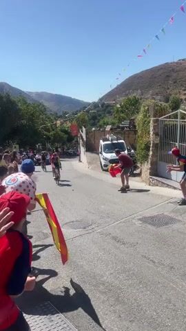 A group of cyclists wearing racing jerseys and helmets are riding up a paved road. The cyclists are mostly male and appear to be in a competition. Spectators are lining the side of the road, some holding Spanish flags and cheering. One man in a red shirt is holding a Spanish flag and appears to be waving it at the cyclists. A white van with police markings is parked on the side of the road. The road is lined with houses and trees, and the background shows a mountainous landscape under a clear blue sky. Decorative colorful flags are strung across the road in the distance. The overall scene depicts a sporting event taking place on a sunny day in a scenic, possibly rural, location.