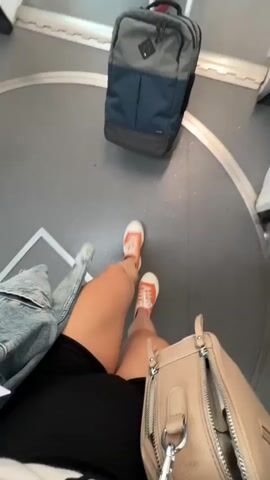 The video begins with a POV shot looking down at a person's legs and feet wearing orange sneakers and black shorts, with a tan purse in the foreground and a blue and gray backpack further back. The perspective shifts to show the person walking through an airport jet bridge. Several other people are walking ahead, each with luggage. The jet bridge has large windows on the right side, offering a view of an airplane parked at the gate and the tarmac. The lighting appears to be artificial from overhead fluorescent lights. The scene is clean and functional, typical of an airport.