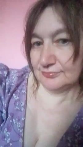 A close-up video shows a woman with shoulder-length brown hair and a purple floral dress with a V-neck. She is looking at the camera and smiling. The background is a pink wall with a portion of a blue and red object visible in the upper right corner. The lighting is soft.