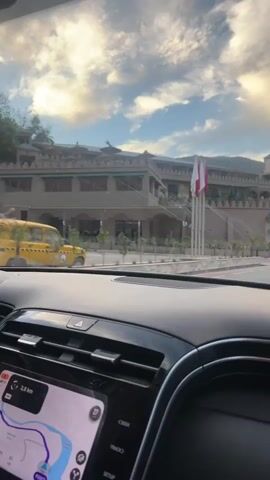 A view through the windshield from inside a car shows the car moving along a road approaching a large building in front. The building has a castle-like appearance with beige walls, a tower, and decorative elements. There are three flags on poles in front of the building. The dashboard of the car is visible, showing the navigation system displaying a map and a distance of 3.8 km. The sky is partially cloudy with the sun shining through.