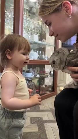 A young child in a white tank top and grey striped pants stands facing a young woman who is holding a grey tabby cat. The child is looking at the cat and the woman is looking at the child. The background shows a wooden cabinet with glass doors displaying various dishes and decorative items. The floor has a patterned tile design. The lighting appears to be indoor ambient light. The scene suggests a moment of interaction between the child, woman, and cat.