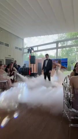 The video opens with a young man in a tuxedo standing in a cloud of fog on a wooden floor. He is looking to his left. A large window is visible behind him, showing trees and sky outside. To his left, a large screen displays a landscape image. In the background, there are speakers and stage lighting. People are seated at tables to the left and right of the frame. The young man turns to his right, and a young woman in a white ball gown walks into the frame and stands next to him. They are both looking at each other and smiling. They then begin to dance in the fog, turning and holding each other. The woman's dress fans out as she turns. The camera zooms in slightly as they continue to dance.