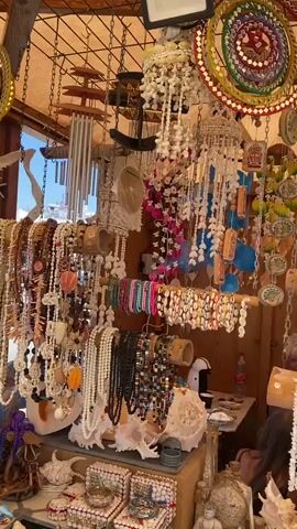 The video shows a busy market stall filled with a wide variety of shell and beaded crafts. In the foreground, there are large conch shells and intricate shell decorations. Above these, numerous strands of necklaces and bracelets made of shells and colorful beads are displayed on wooden racks. Some strands feature white shells, others are adorned with small, multi-colored beads in intricate patterns, and some incorporate larger, polished stones. Hanging from the ceiling are various decorative items, including shell wind chimes and elaborate dreamcatcher-like ornaments made of shells and colorful threads. Small figurines and decorative boxes are also visible on the shelves, adding to the rich, cluttered aesthetic of the stall. The lighting suggests an outdoor setting, possibly under a canopy, casting a warm glow on the merchandise.