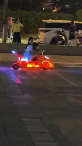At night, two children are riding in a small, brightly-lit red toy car on a paved surface. The toy car has a spoiler and is illuminated with blue, red, and yellow lights. The children are sitting side-by-side, with the child on the right appearing to be steering. In the background, several people are walking on a sidewalk and vehicles, including a white car and a scooter, are visible. There are trees and bushes in the background as well. A person wearing yellow walks by the left side of the shot.