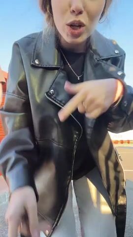 A young woman is standing in what looks like a parking lot, wearing a black leather jacket, light blue jeans, and a black T-shirt. She has reddish-brown hair. She makes hand gestures and lip-syncs to music. The sky is blue. A man walks into the frame and poses with her. He is wearing a gray T-shirt and black pants. He puts his arm around her. There is a small orange structure behind them, and a white semi-truck trailer can be seen in the background. The man appears to be smoking. They both point toward the camera.