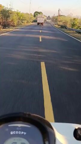 A first-person perspective video captures a view from a scooter traveling on a paved road. The scooter's digital speedometer displays a decreasing speed, starting at 58 km/h, then 57, 52, 47, and finally 44 km/h. The road has a yellow dashed line marking the center and a solid yellow line on the right side. In the distance, two white trucks are visible, with the lead truck appearing to be slightly further ahead. The environment is rural, with sparse vegetation and trees lining the sides of the road. The sun appears to be low in the sky, casting long shadows. The sky is clear and blue. There are power lines visible along the side of the road.