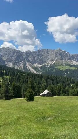 The video pans across a mountainous landscape on a sunny day. The foreground features a lush green meadow with some wildflowers. In the middle ground, a dense forest of dark green coniferous trees stretches across the frame. A small, rustic wooden cabin with a white roof is visible nestled amongst the trees. In the background, a majestic mountain range with rocky, grey peaks and some snow patches rises against a bright blue sky dotted with fluffy white clouds. The camera movement is slow and sweeping, offering a panoramic view of the serene natural environment. There are no people visible in the video.