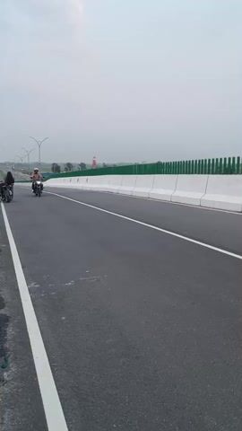 This media file captures a motorcyclist riding on a modern highway in Rupganj, Bangladesh. The scene appears to be a late afternoon or early evening, as indicated by the soft, overcast sky with hints of color that suggest the approaching sunset. The highway is well-maintained, with clear white lane markings and a protective barrier along the side, topped with green fencing.

The primary subject is a person riding a blue and white motorcycle. They are wearing a helmet with a colorful design and a light pink shirt. Their gaze is directed forward as they navigate the road. Another motorcyclist is visible further down the highway, also riding in the same direction.

The setting is an elevated section of the highway, possibly a flyover or bridge, with the landscape visible in the distance. The atmosphere is calm and peaceful, with the open road and the subdued lighting creating a serene mood. No specific event is taking place, but rather a moment of travel and transit.

A license plate is visible on the back of the motorcycle, showing some text and numbers in Bengali script. The overall impression is one of modern infrastructure facilitating movement through a developing region.