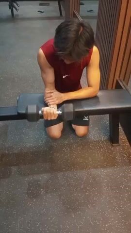 A person is kneeling on the floor of a gym, with their forearms resting on a workout bench. Their right arm is bent at the elbow, and they are holding a dumbbell in their right hand. The dumbbell is being moved up and down by bending and straightening the wrist. The person is wearing a dark red sleeveless shirt and dark shorts. The gym floor is dark with a speckled pattern. The bench is black. Behind the person is a wall with vertical wooden slats.