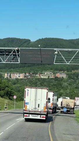 A digital sign is mounted on an overhead gantry above a road. The sign displays text in Russian, warning about reversible traffic at kilometer 1766. The text appears to be "Внимание!!! на км 1766 (Атлян) Реверсивное движение". The sign is illuminated and readable. Below the sign, a road with multiple lanes is visible, with several trucks driving on it. The environment is sunny with a clear blue sky and a backdrop of lush green forested hills and some buildings. A person appears to be working on or near the sign on the left side of the gantry, possibly maintenance or installation.