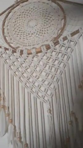 The video shows a close-up of a dreamcatcher hanging on a white wall. The dreamcatcher has two hoops. The top hoop has a white crocheted center. The bottom hoop has a woven, diamond-patterned macrame design. Long white strands hang down from the bottom hoop, adorned with wooden beads and white feathers. The camera moves slightly during the video.