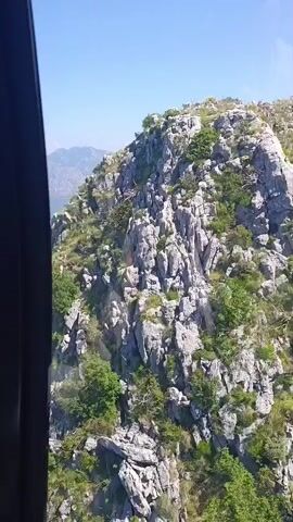 This is a video captured from a moving vehicle, likely a car or bus, looking out of a window at a mountainous landscape. The scene is dominated by rugged, rocky terrain covered with sparse green vegetation. The mountains are layered, with the foreground featuring sharp, grey rock formations and scattered trees. In the distance, a vast expanse of hazy blue mountains meets a clear, bright blue sky.

The activity appears to be a scenic drive or journey through the mountains. The camera's perspective suggests movement, as the view subtly shifts. The time of day is clearly daytime, indicated by the bright sunlight and the shadows cast on the rocks. The weather is fair and sunny, with no clouds visible in the sky.

The location is identified as Škaljari, Montenegro. This context suggests the view might be from a road winding up or down a mountain in that region, offering panoramic views of the surrounding natural beauty. The rocky, steep terrain and the layered mountain ranges are characteristic of the Montenegrin landscape. There are no people or specific objects visible that would indicate a particular event, beyond the journey itself. The overall mood conveyed is one of awe and appreciation for the natural scenery. There is no visible text in the media file.