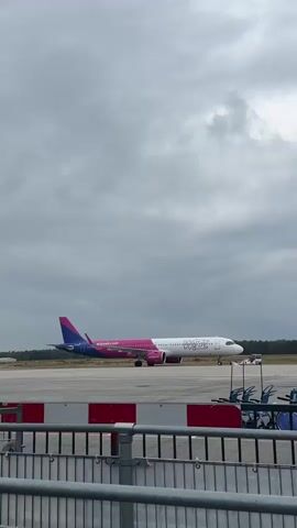 A Wizz Air airplane is seen on a tarmac under a cloudy sky. The plane is white with pink and purple accents on its tail and rear fuselage. It is positioned on a runway with other vehicles and structures visible in the background. A gray metal fence is in the foreground.