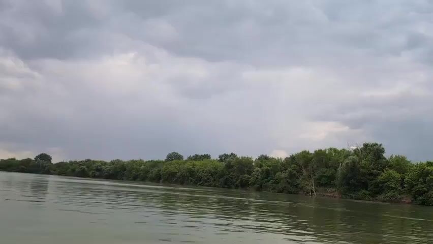 The video shows a wide, calm river with murky green water. The riverbanks are lined with dense, lush green trees. The sky is overcast with dramatic, dark grey clouds. The perspective suggests the viewer is on a boat moving slowly down the river. There are no people or animals visible in the frame. The overall atmosphere is serene but also hints at the possibility of impending rain due to the heavy clouds.