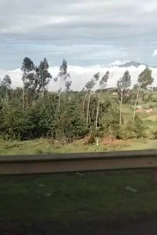 The video shows a scenic view of a green landscape with trees and fields, possibly from a moving vehicle. The camera is positioned at a slight angle, looking out over a railing. In the background, there is a mountain or hill partially obscured by clouds. The sky is filled with a mix of blue and white clouds. The video pans slightly to the side, showing more of the landscape and the cloudy sky. The focus seems to be on capturing the natural beauty of the scenery, with the presence of trees, fields, clouds, and distant mountain being the main elements.