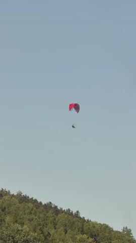 The video shows a person paragliding against a clear sky. The paraglider is red and is suspended high in the air. Below the paraglider, a lush green forest can be seen. The video is taken from a distance, giving a sense of the vastness of the sky and the height of the paraglider. The sun appears to be shining, as indicated by the bright sky and the clear visibility of the scene. The paraglider seems to be moving slowly across the sky.