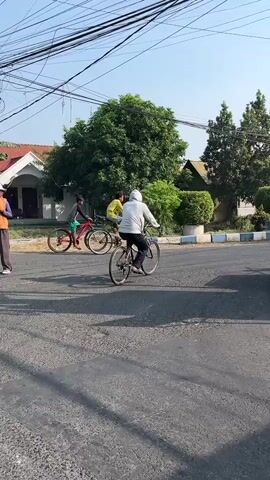The video shows an outdoor scene on a paved road with several people riding bicycles and motorcycles. In the foreground, a person wearing an orange vest and a white helmet stands in the middle of the road, holding a red flag and a yellow flag. This person appears to be directing traffic or signaling. Behind them, three people on bicycles are visible, one in the foreground and two further back. Further down the road, several motorcycles are also present, some in motion and some parked. Trees and buildings line the side of the road, and overhead power lines are prominent in the frame. The sky is clear and appears to be daytime. The overall impression is of a busy street with various modes of transportation and traffic control.
