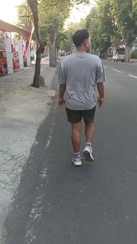 A person walks away from the camera down a paved road. The person is wearing a grey t-shirt, dark shorts, and athletic shoes. Trees line the left side of the road, and buildings and other trees are visible in the background on the right. The sun appears to be setting, casting long shadows.