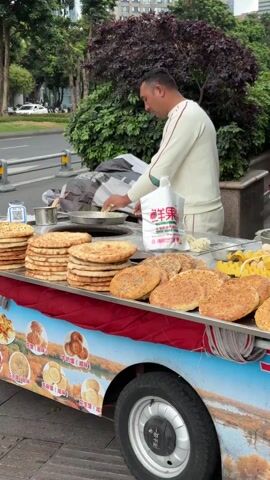 A man is preparing food at a street food cart. He is wearing a white top and pants with red trim. There are several stacks of flatbread on the cart, some plain and some with seeds. The cart also has eggs and other ingredients. The man is working on dough over a round open oven in the top of the cart. The cart itself is decorated with a picture of a landscape. The background shows a street with trees and cars.
