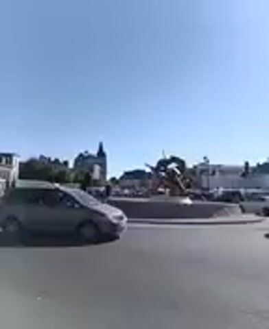 A video captured from a vehicle shows a view of a roundabout with a sculpture in the center. Several cars are seen driving around the roundabout and on the surrounding roads. The sky is clear and blue. Buildings are visible in the background. The video is blurry due to motion.