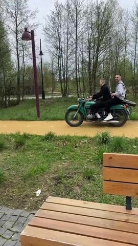Two people are riding a green motorcycle with a sidecar down a yellow path. A person on a bicycle passes them in the opposite direction. The motorcycle continues down the path, passing benches and trees. Other people are visible in the background, sitting on a bench. The scene takes place in a park-like setting. The weather appears overcast.
