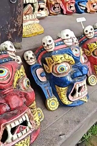 The video shows a display of colorful, carved wooden masks, many with skull motifs. The masks are arranged on wooden shelves, with some in the foreground and others further back. The masks are intricately designed with bold colors like blue, red, yellow, and white. Many of them have exaggerated facial features, including wide-open mouths with sharp teeth, bulging eyes, and fierce expressions. The masks appear to be decorative or possibly for ceremonial purposes, and they are displayed in what seems to be an outdoor market or shop setting. The lighting suggests it is daytime. There are no people visible in the immediate frame, but the context suggests a vendor is present.