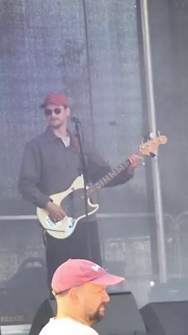 The video captures a brief scene on what appears to be a stage. In the foreground, the back of a person's head wearing a pink baseball cap is visible. Behind them, a man wearing a grey button-up shirt, black pants, sunglasses, and a red baseball cap is playing a light-colored bass guitar. He is positioned in front of a large, textured grey backdrop. To the left of the bass player, another man, wearing a light-colored short-sleeved shirt and dark pants, is also on stage. He is facing away from the camera, holding a black cable in his left hand and appears to be interacting with some equipment. The lighting suggests an outdoor or large indoor venue, possibly a concert or performance.
