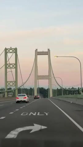 A drone shot captured the sunset with a pink and white sky above a suspension bridge, illuminated by an orange street light. The camera then shifted to an eye-level perspective from the driver's seat of a car driving on the bridge. The car's perspective showed the road ahead, with a white BMW in front. The bridge had two green towers, each supporting suspension cables that stretched across the road. The sky was a soft pink and white, indicating either sunrise or sunset. The bridge was wide, with multiple lanes of traffic, and trees lined the background. The video captured a serene moment as the sun set, casting a warm glow over the bridge and the surrounding landscape.