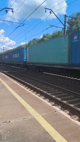 A train carrying shipping containers travels along a railway track. The containers are of various colors, including blue, green, and red. Some of the blue containers have the word "SINOKOR" printed on them in large white letters. The train is passing a railway platform with a yellow line marking the edge. The sky is blue with some white clouds. Trees are visible in the background. Overhead wires for the electric train are also visible.