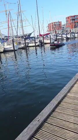 A small inflatable boat with two people on board navigates through a marina filled with sailboats. The boat is dark in color with red trim. The water is blue and calm, with reflections of the boats and buildings. The marina has a wooden dock in the foreground. In the background, there are several tall sailboats with masts and sails, and modern buildings with red brick facades. The sky is clear and blue.