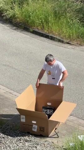 In a sunny, outdoor setting, a middle-aged man wearing a white t-shirt, dark shorts, and sandals opens a large cardboard box on the edge of a paved driveway. The box contains a robotic lawnmower and a black and white charging station. The man removes the cardboard flaps of the box and then a long, narrow cardboard piece. A patch of overgrown grass is visible behind him. The ground is a mix of gravel and pavement.