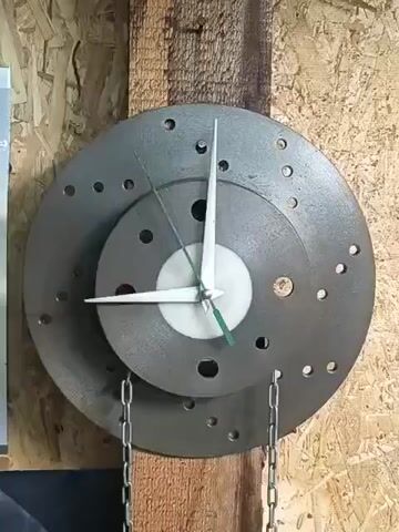 The video shows a close-up of a handmade clock mounted on a wooden beam. The clock itself is constructed from two stacked brake rotors, the larger one underneath and a smaller one on top. The face of the clock has no numbers. Three hands are visible: a long, thin hour hand, a shorter minute hand, and a thin second hand. The hour and minute hands are white, while the second hand is green. Two silver chains hang down from the bottom of the clock, one on each side. The background appears to be a wall made of wood panels.