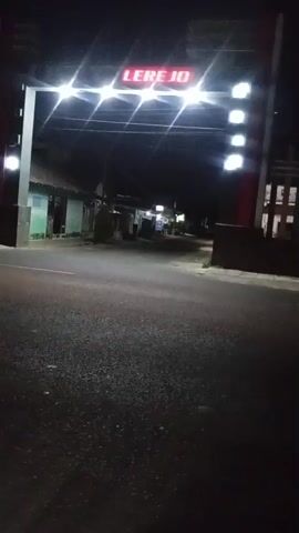 A dark, paved road is visible at night, with a large archway spanning across it. The archway has bright, white lights illuminating it, and a red digital sign above reads "LEREJO". On the left side of the road, a car with its headlights on is partially visible, moving away from the camera. The background shows dark silhouettes of buildings and trees, suggesting a rural or suburban setting. The overall scene is dimly lit, with the bright lights of the archway and the car's headlights being the primary light sources.