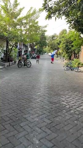 The video shows a street scene in Kalisampurno, Indonesia. The street is paved with interlocking bricks and lined with houses and trees. Several people, mostly children, are present. Two boys are on bicycles on the left side of the street, and another boy is standing in the middle of the street holding a gray helmet. Further down the street, a boy in a blue shirt and red shorts is holding a blue and white umbrella, seemingly playing with it. Another person, possibly a boy, is walking away from the camera in the background. The scene appears to be set during the day, possibly late afternoon or early evening, given the lighting. The overall atmosphere seems playful and casual, with children engaged in outdoor activities. There are no other vehicles in motion, and the general environment suggests a residential area. No text is clearly visible in the video.