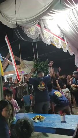A group of people are gathered under a white tent, possibly at an outdoor event. In the foreground, a blue table holds a plate of food items and small cups. In the middle ground, two men in dark blue t-shirts with logos are dancing with their arms raised. One man is facing away from the camera, while the other is partially visible in profile. Other individuals are visible in the background, some with their arms raised, suggesting a lively atmosphere. A flag, possibly a national flag, is visible on the left side of the frame. The lighting appears to be artificial, with bright lights illuminating the tent from above. The overall scene depicts a celebratory or festive gathering.