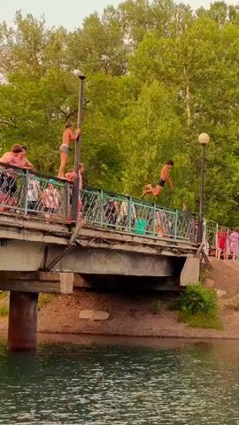 The media file captures a summertime scene at a body of water, possibly a river or lake, near Abakan, Russia. The water is a tranquil blue-green, with visible ripples on its surface. On the bank, a group of people are gathered. In the foreground, a young boy in swim trunks stands in the shallow water. Further back, a slight incline leads up to a bridge.

The main activity depicted is a child jumping off the side of the bridge into the water. The moment of impact creates a splash, captured in the second frame. On the bridge and the bank, several other individuals are visible, some walking and others standing. They appear to be casually dressed, suggesting a warm day. The background is dominated by lush green trees, indicating a verdant environment.

Notable details include the presence of a lamppost on the bridge and a metal railing. The lighting suggests it is daytime, possibly late afternoon or early evening given the soft, diffused light. The mood appears relaxed and recreational. There is no visible text in the media file. The overall scene conveys a sense of summer fun and outdoor activity.