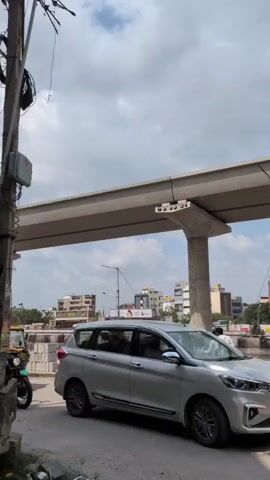 The video displays a daytime urban street scene under a partially cloudy sky. An elevated concrete structure, consistent with a metro or monorail track, spans across the frame, supported by a large pillar.

In the foreground, a person wearing a helmet and a dark shirt rides a dark-colored motorcycle (likely a Royal Enfield Scram 411) from left to right. Following the motorcycle, a green and yellow auto-rickshaw, driven by a person, also moves from left to right. A silver multi-purpose vehicle (MPV), possibly a Maruti Suzuki Ertiga, is partially visible on the right side of the frame, either parked or moving slowly in traffic.

Utility poles with various wires are seen on the left side. In the background, beyond the road, several multi-story buildings are visible, some appearing to be under construction with scaffolding. The word "EVEREST" is visible on a billboard or sign on the left side, behind where the auto-rickshaw passes. The general setting and vehicles are indicative of a city environment in India, specifically Bengaluru as per the context provided. There are no discernible emotions displayed by the individuals.