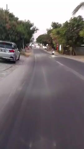 The video shows a view from a moving vehicle traveling on a paved road. On the left side of the road, there is a sidewalk with some vegetation and a few small buildings or stalls. A grey car is parked on the left side of the road. The road itself is mostly empty, with a few other vehicles visible in the distance, including a motorcycle with a rider. The sky is overcast. The overall impression is that of a rural or suburban road in daytime.