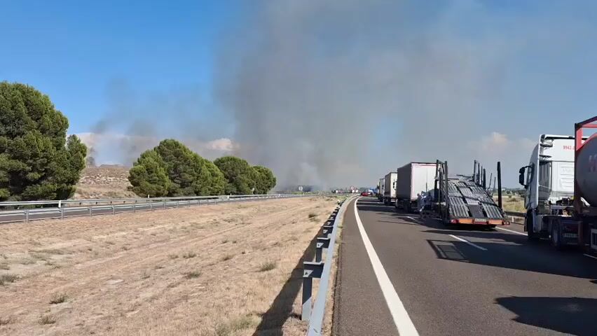 The video shows a traffic jam on a highway. A large plume of dark smoke is visible in the distance, suggesting a fire. Several trucks are stopped on the road, facing away from the camera. Some of the trucks are carrying large containers. The sky is clear and blue, and the sun is shining. The area beside the highway appears to be dry and grassy, with some trees in the background. There is a guardrail separating the highway from the roadside.