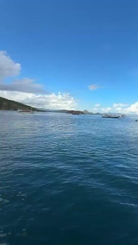 A short video shows a small, red motorboat in the middle of the ocean carrying several people. A shirtless man stands at the front of the boat, while the rest of the passengers are seated. In the background are green islands and other boats, including a sailboat. The sky transitions from blue with white clouds to overcast with darker clouds. The water is deep blue with gentle ripples. The video is shot from another boat, with rigging visible in the foreground in the final frame.
