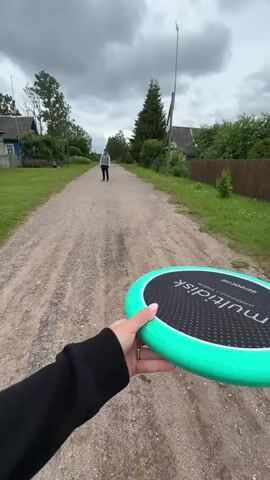 The video shows a person holding a green frisbee with a black center and white text that appears to say "ULTRA DISK". The frisbee is in the foreground, partially obscuring the rest of the scene. The camera is moving forward along a dirt road in what appears to be a rural setting.

In the background, a person is walking away from the camera down the dirt road. To the left of the road is a grassy field with a weathered wooden fence. Beyond the fence, there are trees and a small, rustic house with a blue accent. A tall, thin pole with some sort of equipment at the top stands behind the house.

The sky is overcast with heavy, gray clouds, suggesting a cloudy or potentially rainy day. The overall atmosphere is somewhat somber due to the weather and the rustic appearance of the location. The presence of the frisbee implies an intention for outdoor recreation, though the weather may be a deterrent. The location appears to be a village in Borki, Russia, given the context provided.
