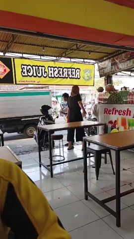 The video captures the view from inside a juice shop looking out onto a street. A yellow banner with the words "REFRESH JUICE" printed in reverse is hanging above the entrance. A person wearing a black shirt and a hijab stands at a table, facing away from the camera. To their left, a young person in a blue shirt and red shorts walks past. A motorcycle is parked near the entrance. On the street outside, there is traffic, including cars and motorcycles. Inside the shop, tables and chairs are visible. The floor is tiled.