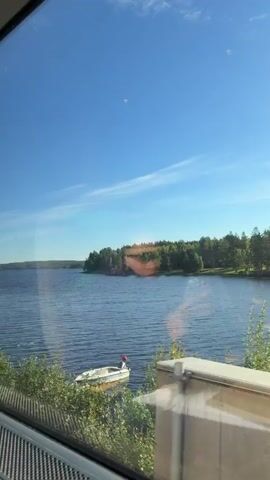 The video shows a view from a train window looking out at a large body of water, likely a lake, under a clear blue sky with a few wispy clouds. Lush green trees and vegetation line the shore of the distant landmass. The water is rippling, suggesting a gentle breeze. There are some reflections on the train window, one of which appears to be the interior of the train. The bottom of the frame shows the edge of the train window structure, with a textured grey material visible. The overall impression is a peaceful, scenic journey through a natural landscape.